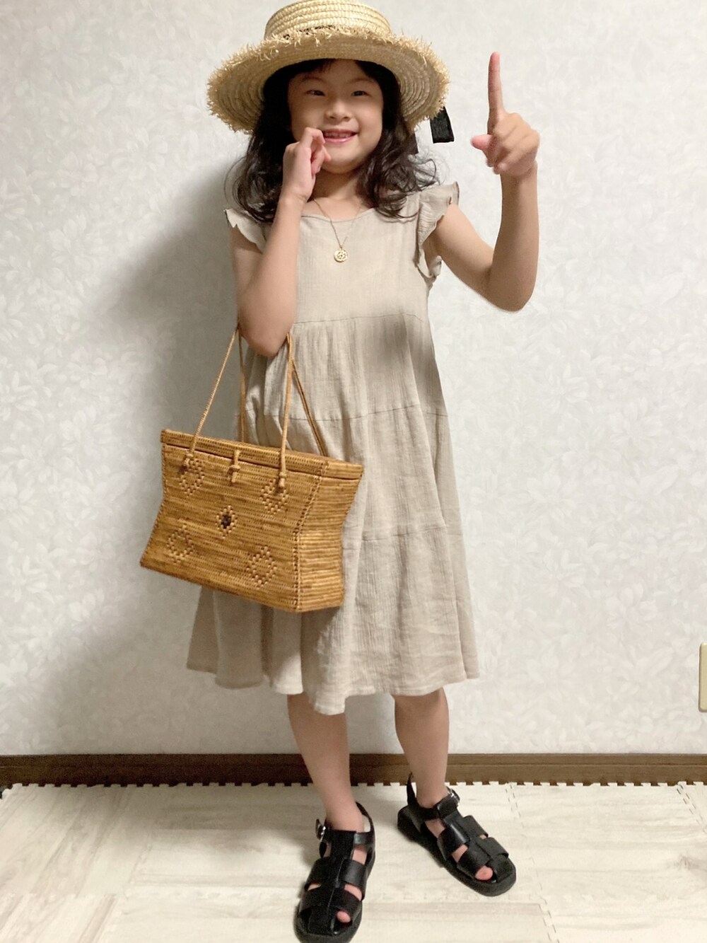 ベージュ系のワンピース、ブラウン系のかごバッグ、ブラック系のサンダルを着用したキッズの夏コーディネートの2枚目の写真