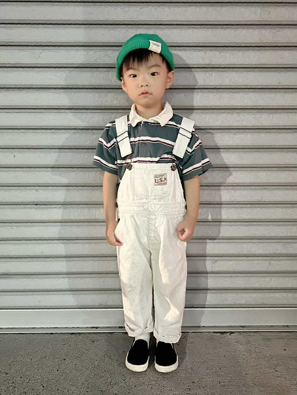グリーン系のポロシャツ、ホワイト系のサロペット/オーバーオール、ブラック系のスリッポンを着用したキッズの夏コーディネートの2枚目の写真