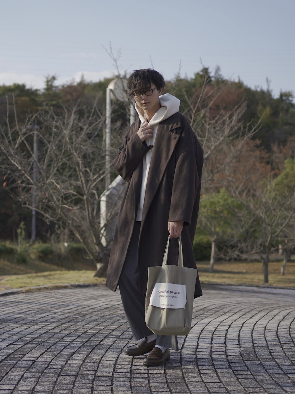 ベージュ系のトートバッグ、ブラウン系のトレンチコート、ホワイト系のパーカーを着用したメンズの秋コーディネートの2枚目の写真
