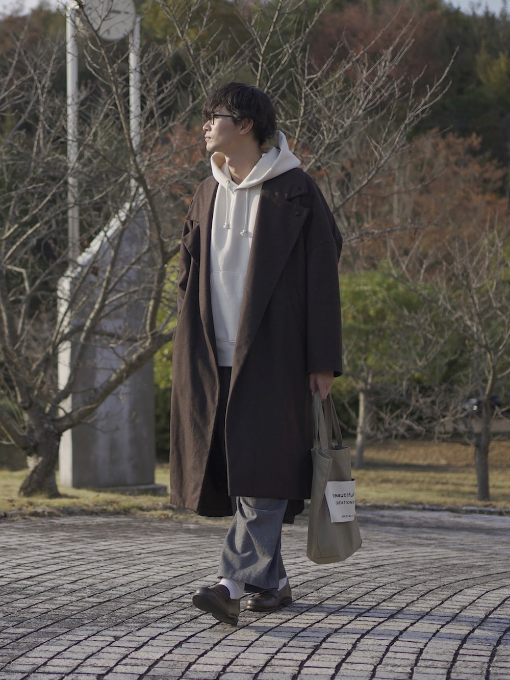 ベージュ系のトートバッグ、ブラウン系のトレンチコート、ホワイト系のパーカーを着用したメンズの秋コーディネートの3枚目の写真