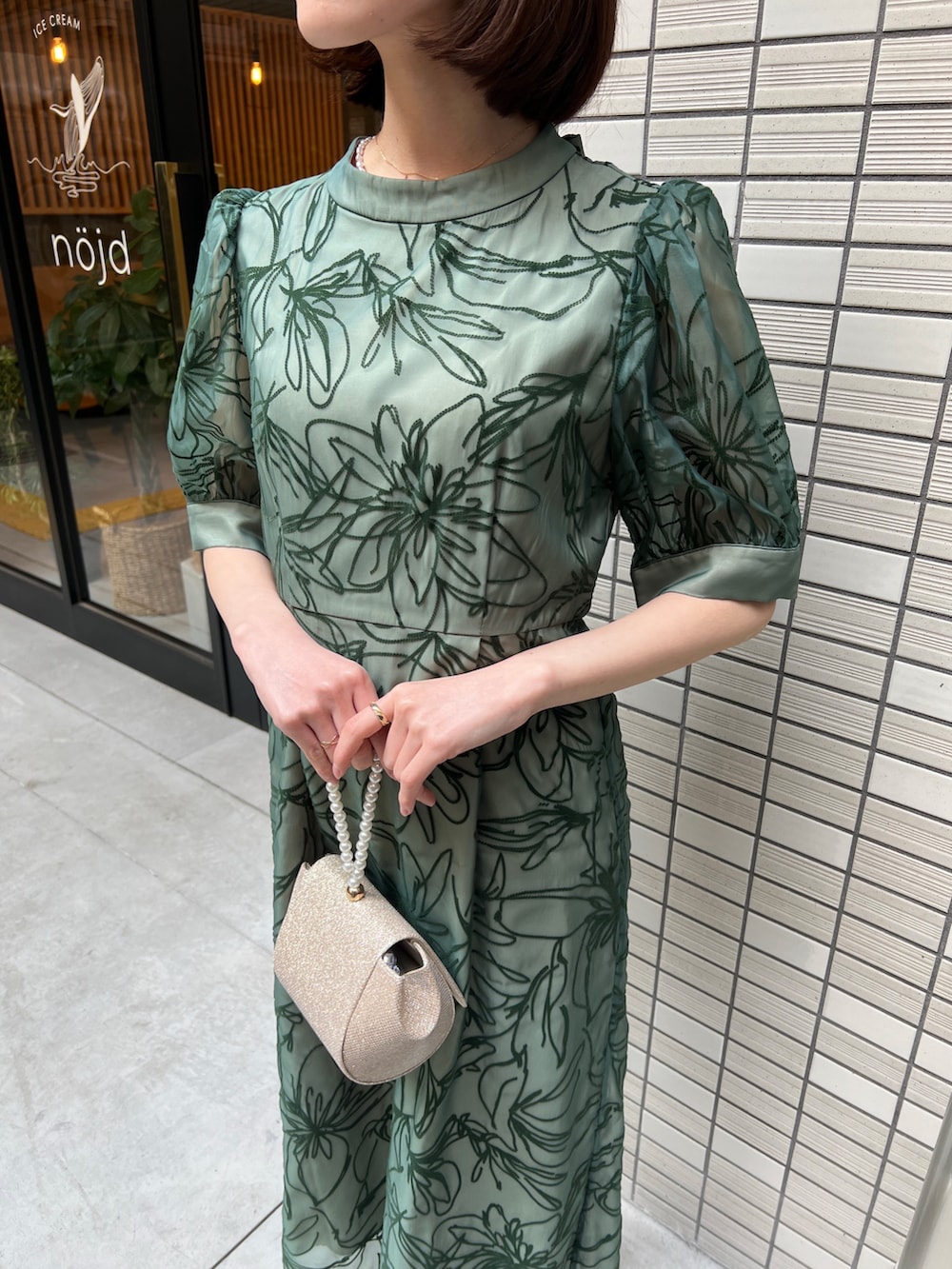 ゴールド系のハンドバッグ、ベージュ系のパンプス、グリーン系のドレスを着用したレディースの春コーディネートの3枚目の写真