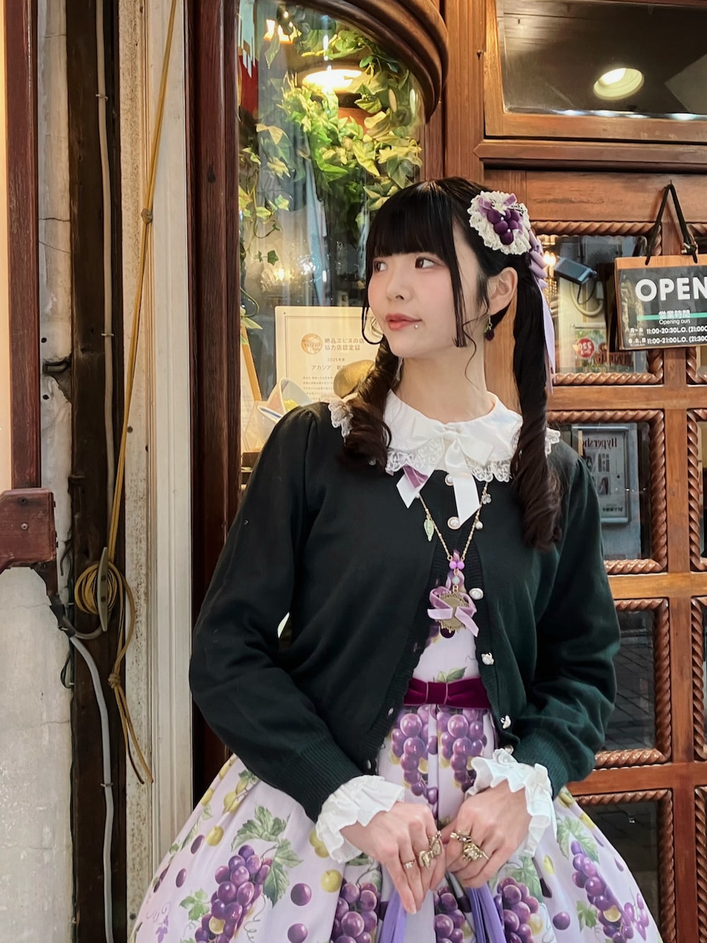 グリーン系のカーディガン/ボレロ、ホワイト系のシャツ/ブラウス、パープル系のジャンパースカートを着用したレディースの春コーディネートの2枚目の写真