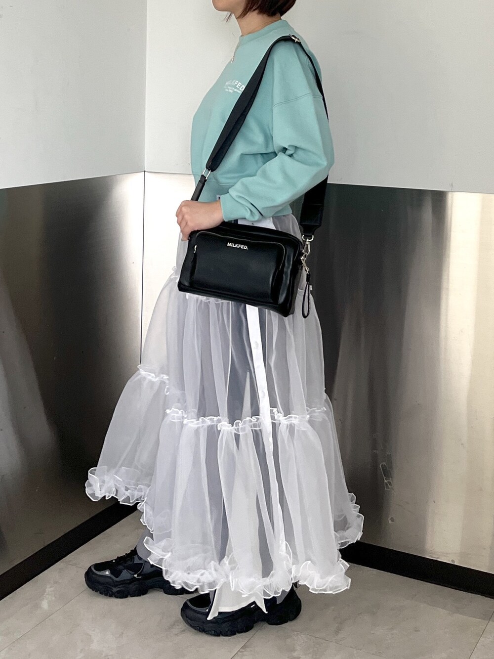 グリーン系のスウェット、ブラック系のショルダーバッグ、ホワイト系のスカートを着用したレディースの春コーディネートの2枚目の写真