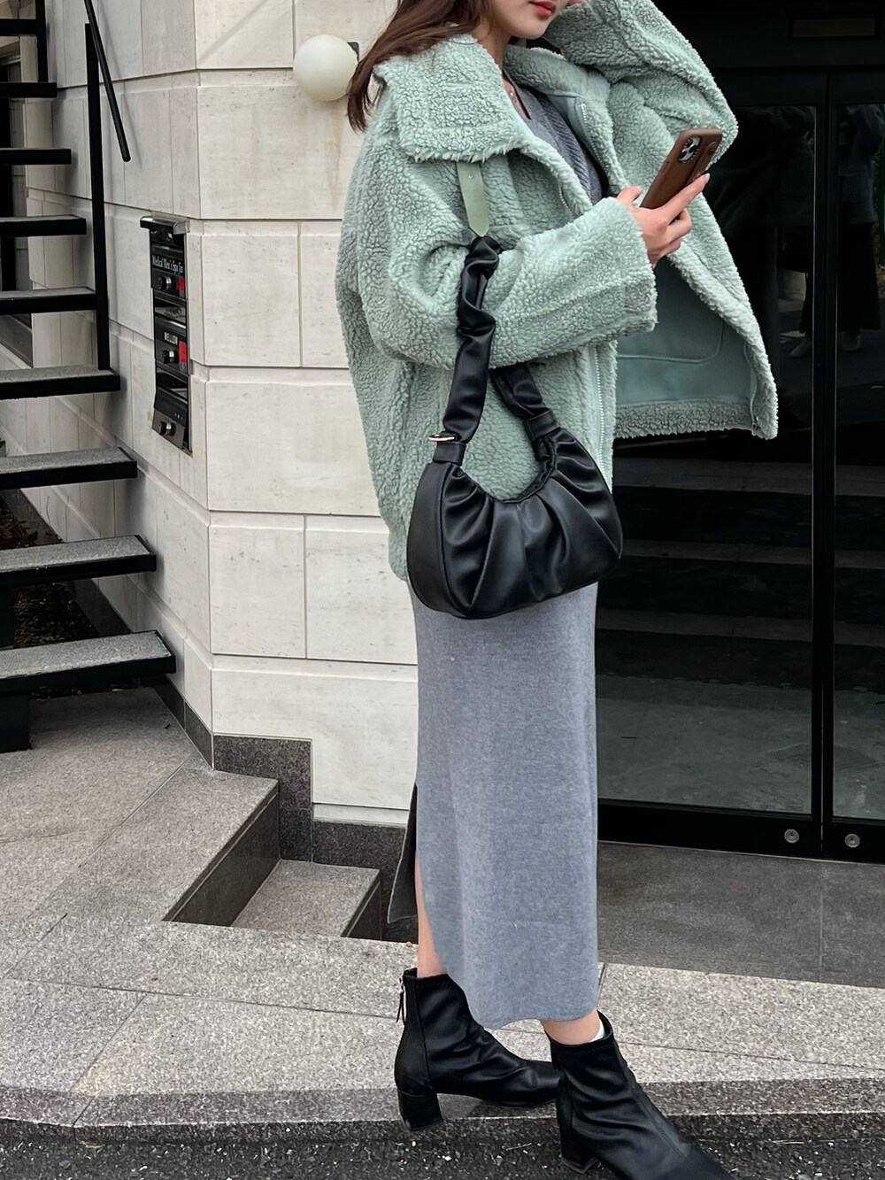 グリーン系のムートンコート、グレー系のニット/セーター、ブラック系のハンドバッグを着用したレディースの秋コーディネートの2枚目の写真