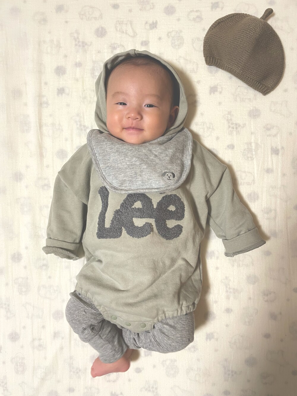 グリーン系のロンパース、グレー系のスタイ/よだれかけ、グリーン系の帽子を着用したキッズの冬コーディネートの3枚目の写真