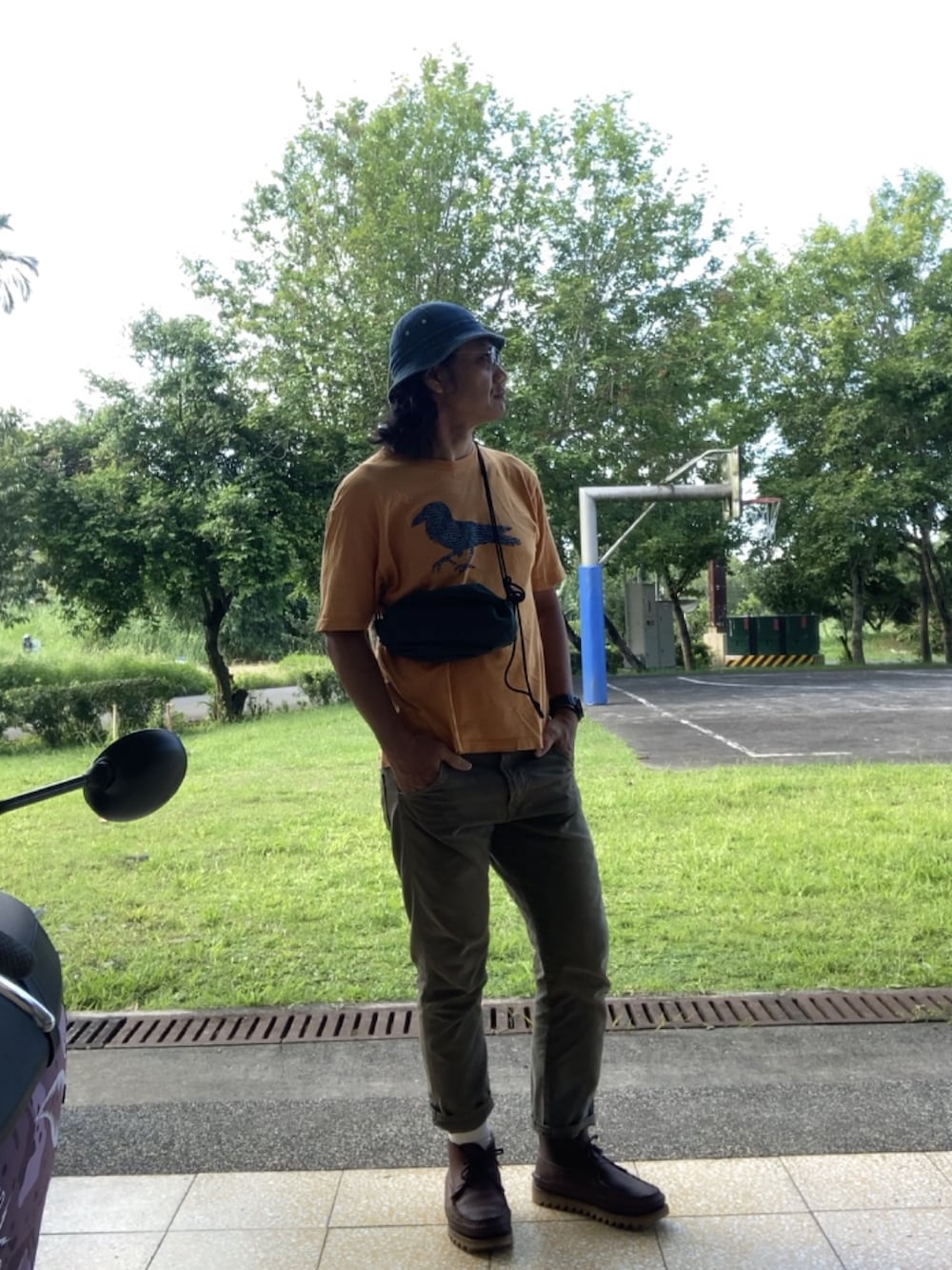 グリーン系のショルダーバッグ、オレンジ系のTシャツ/カットソー、ブラウン系のブーツを着用したメンズの夏コーディネートの3枚目の写真