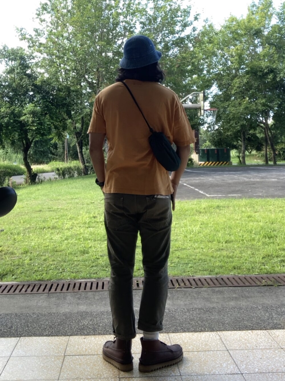 グリーン系のショルダーバッグ、オレンジ系のTシャツ/カットソー、ブラウン系のブーツを着用したメンズの夏コーディネートの2枚目の写真