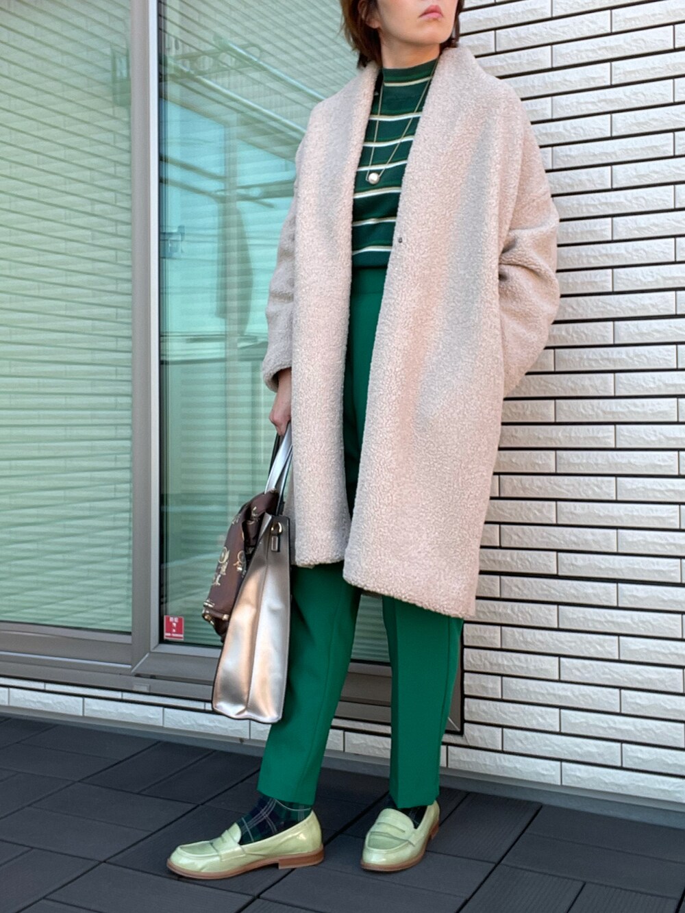 ベージュ系のノーカラーコート、グリーン系のニット/セーター、シルバー系のトートバッグを着用したレディースの冬コーディネートの2枚目の写真