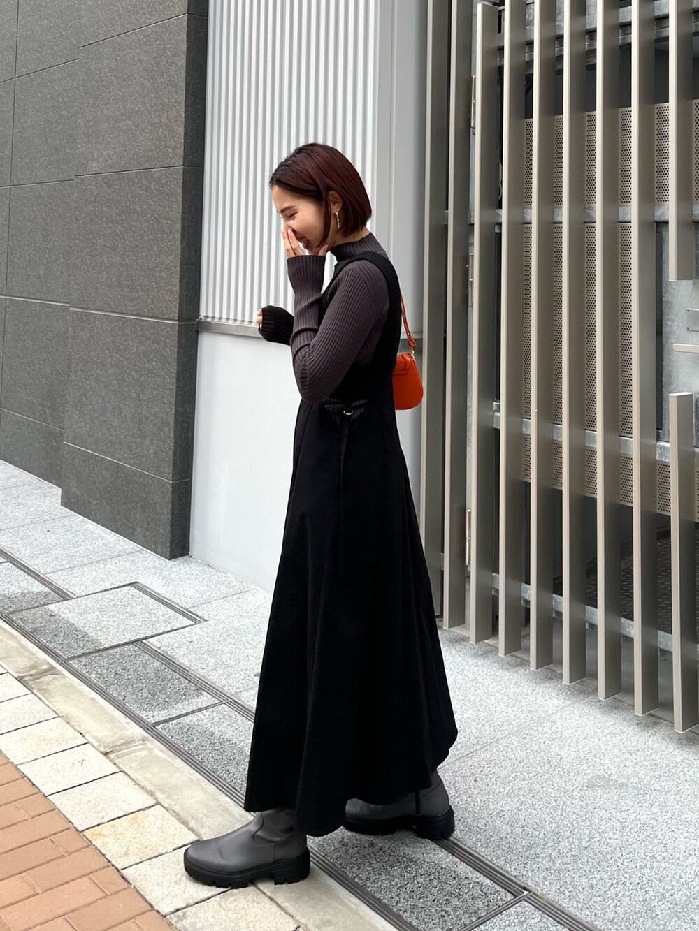 グレー系のニット/セーター、ブラック系のジャンパースカート、オレンジ系のショルダーバッグを着用したレディースの冬コーディネートの2枚目の写真