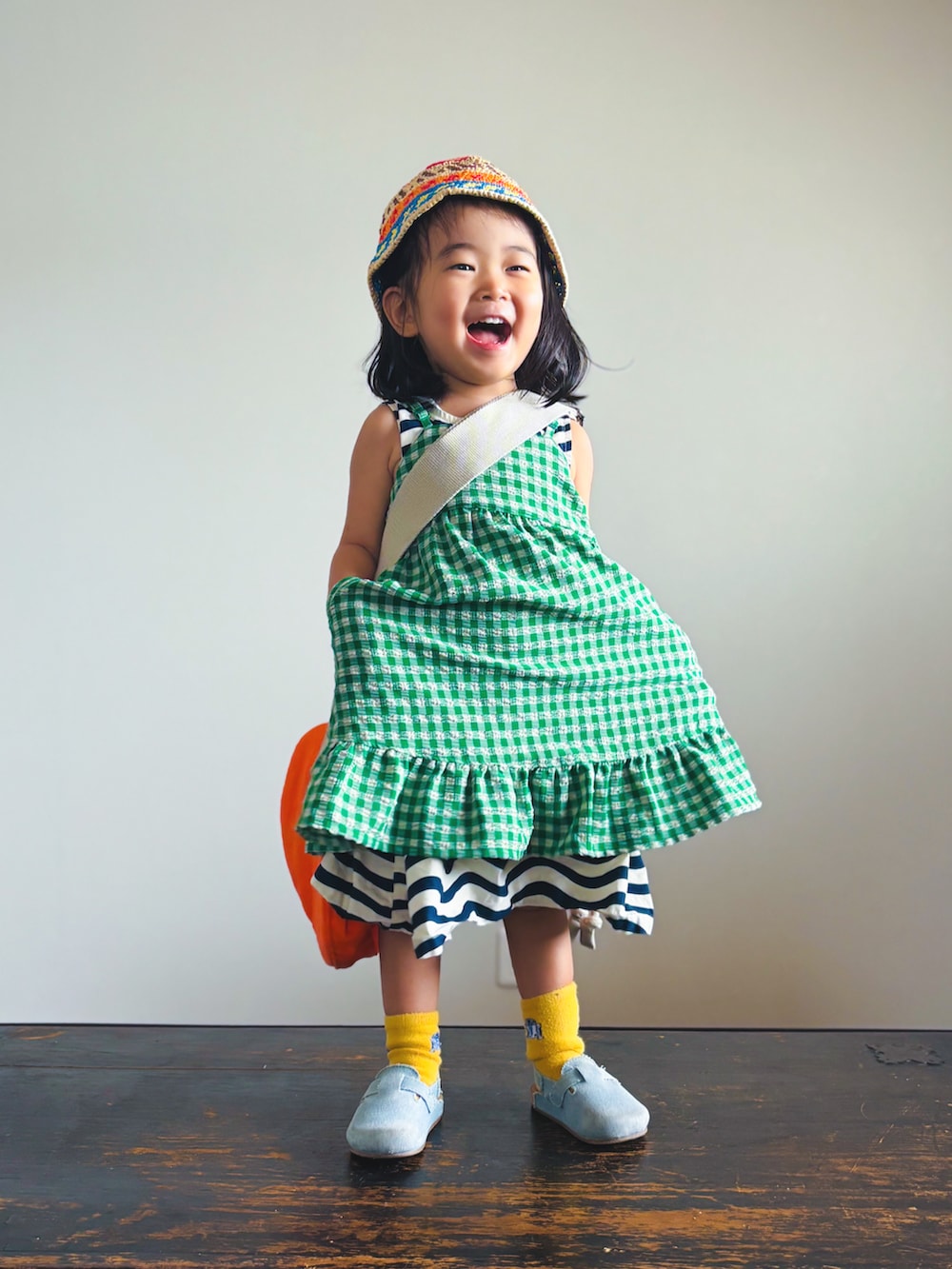 グリーン系のスカート、ブラック系のワンピース、オレンジ系のショルダーバッグを着用したキッズの夏コーディネートの2枚目の写真