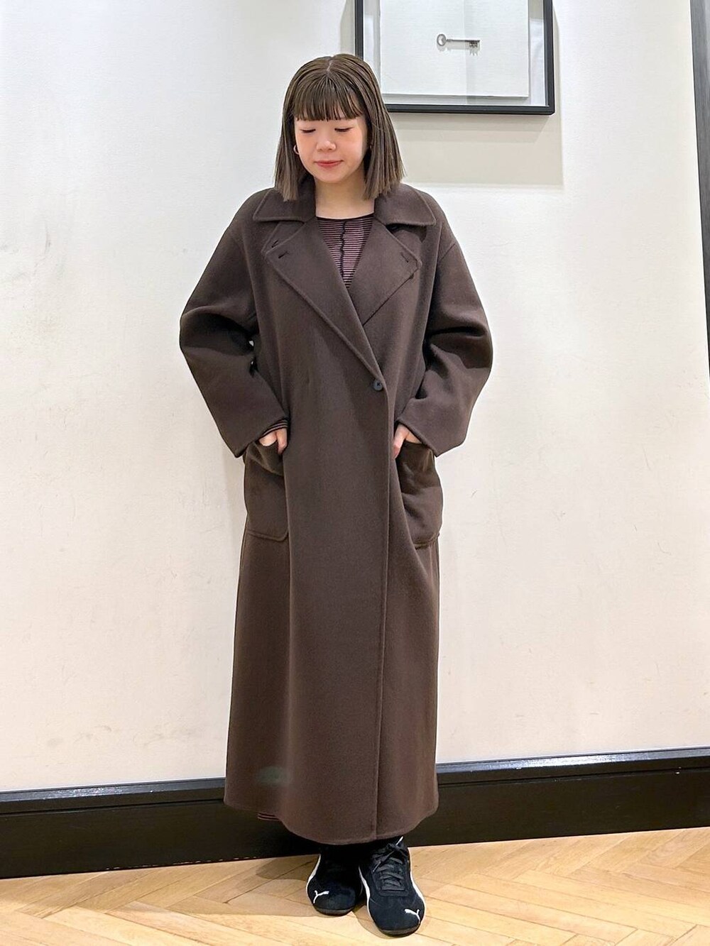 ブラウン系のステンカラーコート、ブラック系のスニーカー、ブラウン系のワンピースを着用したレディースの秋コーディネートの3枚目の写真