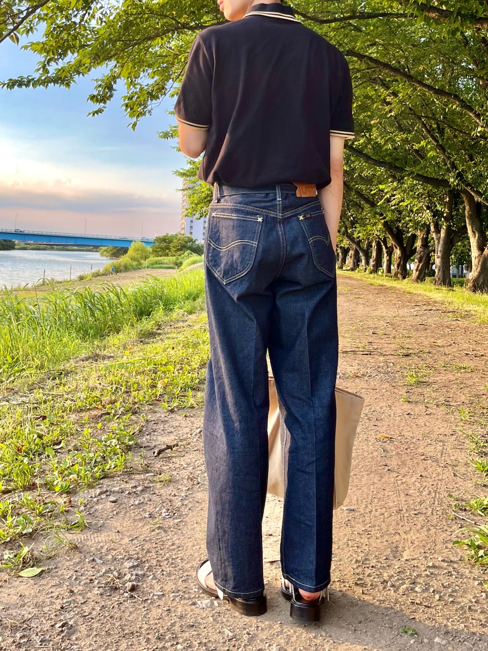 ブラック系のポロシャツ、ブルー系のデニムパンツ、ホワイト系のトートバッグを着用したメンズの夏コーディネートの2枚目の写真