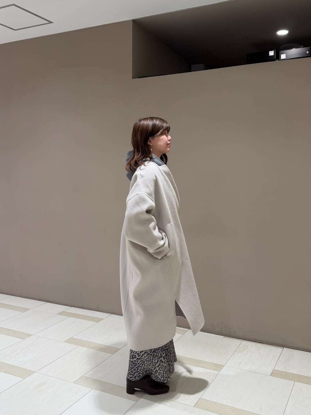 グレー系のパーカー、ベージュ系のチェスターコート、ブラウン系のスカートを着用したレディースの秋コーディネートの2枚目の写真