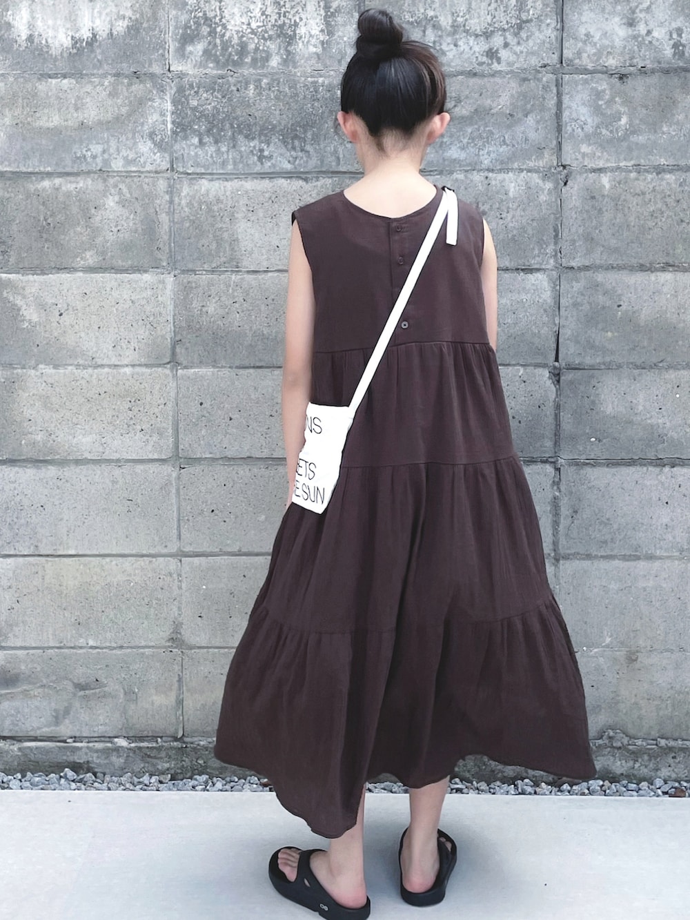 ブラウン系のワンピース、ホワイト系のショルダーバッグ、ブラック系のサンダルを着用したキッズの夏コーディネートの2枚目の写真