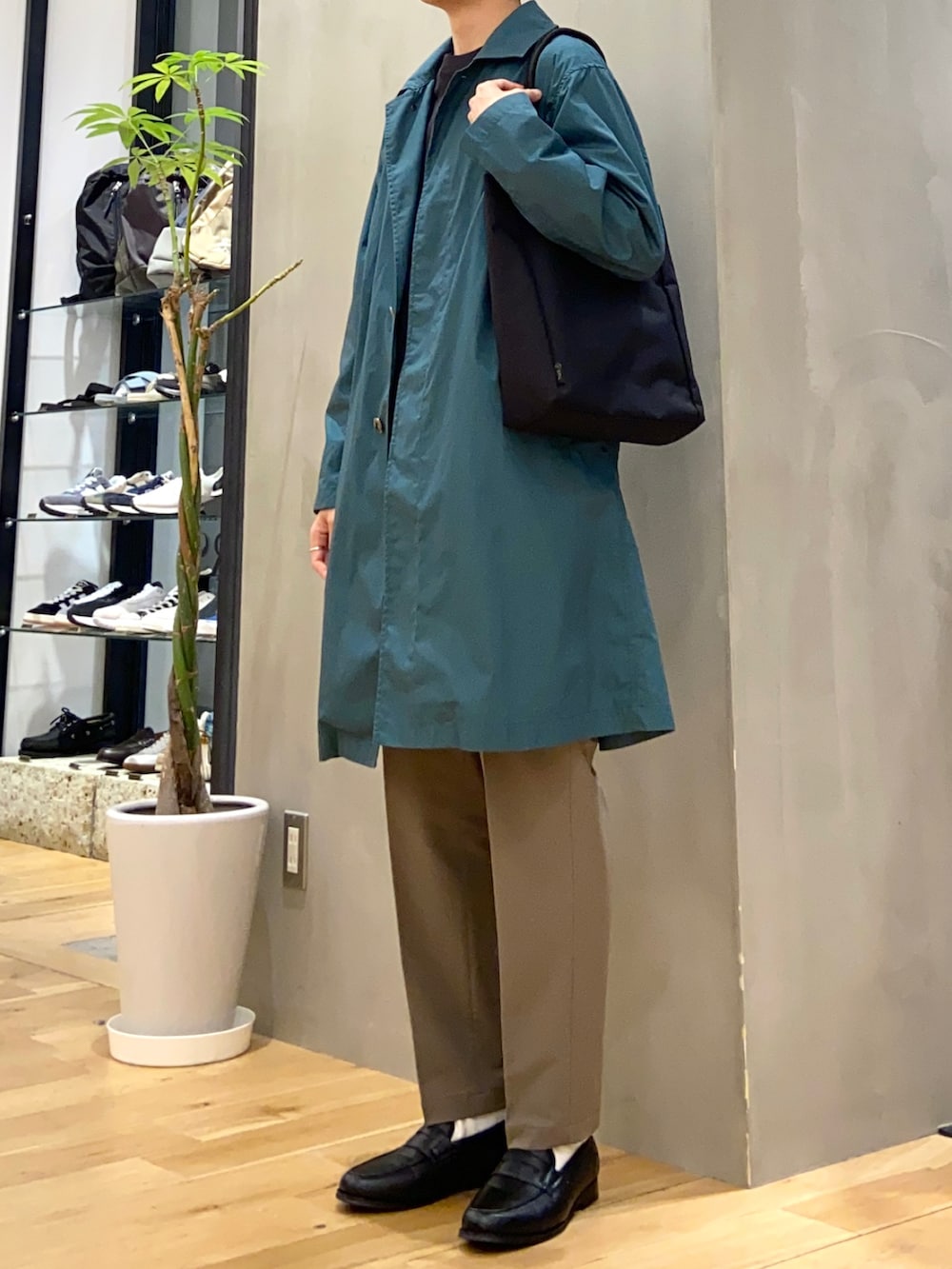 グリーン系のステンカラーコート、グレー系のTシャツ/カットソー、ブラック系のトートバッグを着用したメンズの春コーディネートの2枚目の写真