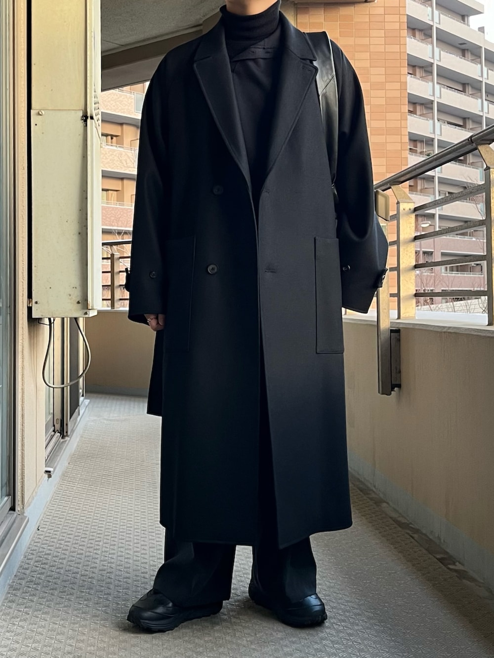 ブラック系のチェスターコート、ブルー系のニット/セーター、ブラック系のショルダーバッグを着用したメンズの冬コーディネートの2枚目の写真