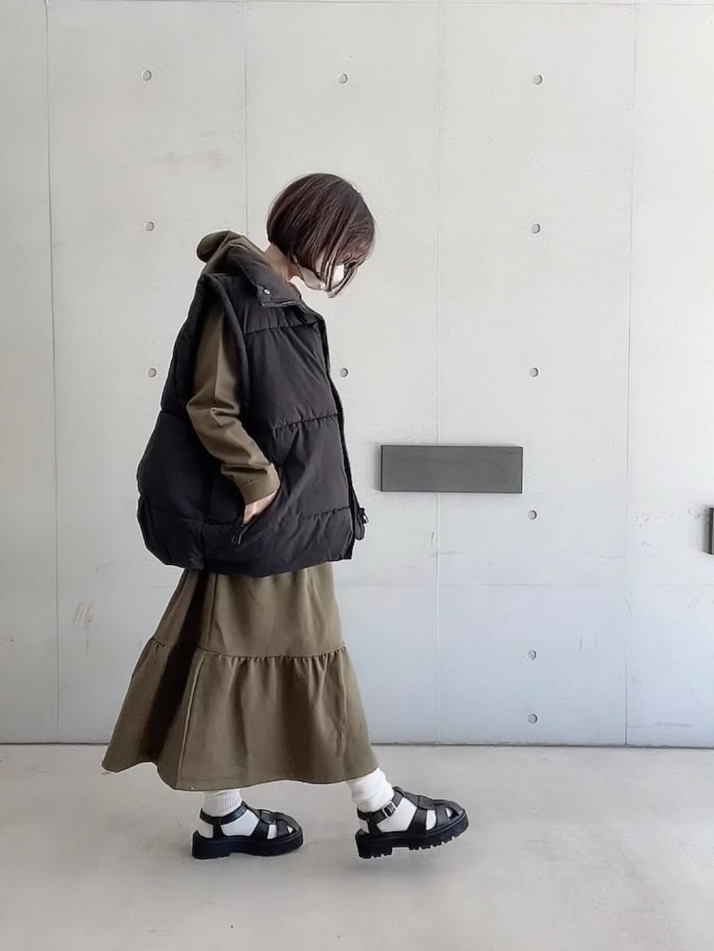 グリーン系のワンピース、ブラック系のダウンベスト、ブラック系のサンダルを着用したレディースの春コーディネートの2枚目の写真