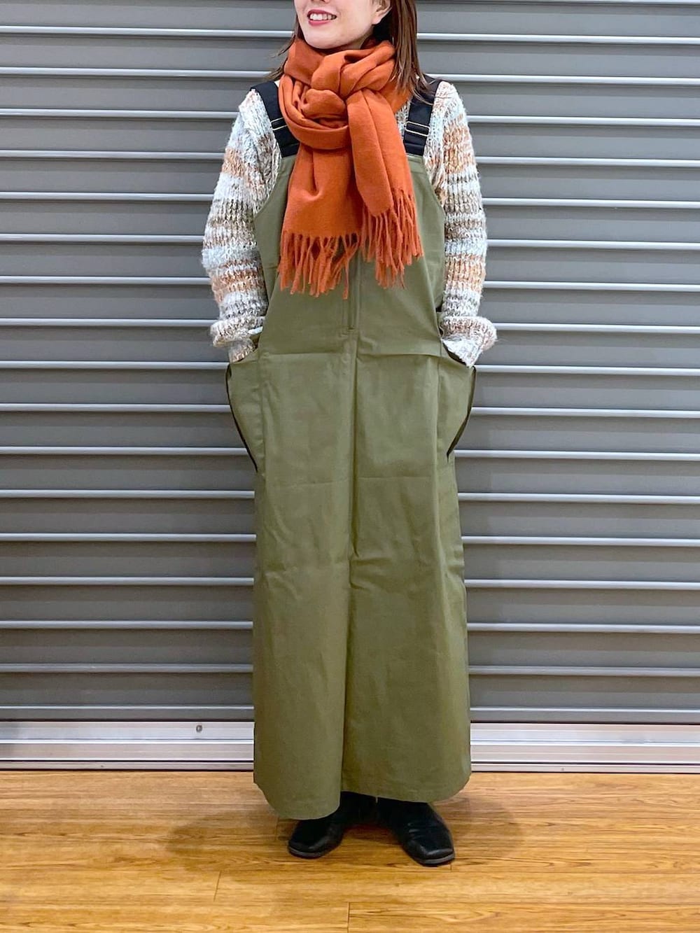 グリーン系のジャンパースカート、ベージュ系のニット/セーター、オレンジ系のストール/ショールを着用したレディースの冬コーディネートの2枚目の写真