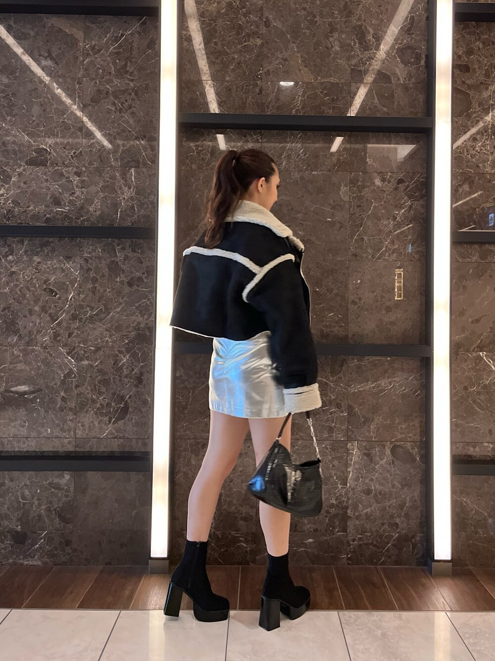 ブラック系のムートンコート、ブラック系のニット/セーター、シルバー系のスカートを着用したレディースの秋コーディネートの2枚目の写真