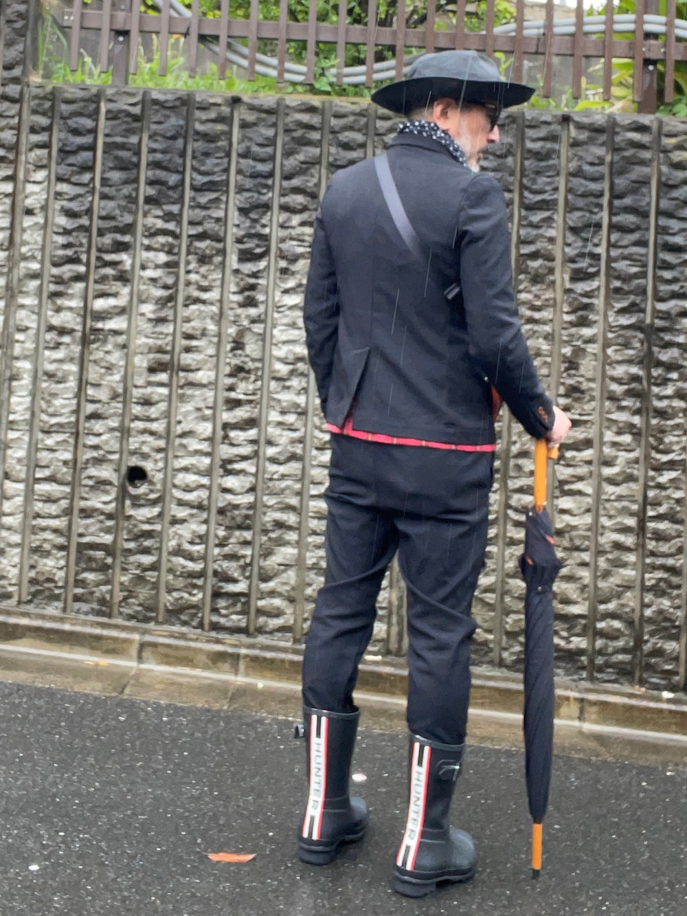 ブラック系のレインシューズ、ブラック系のハット、ブラック系のメガネを着用したメンズの春コーディネートの2枚目の写真