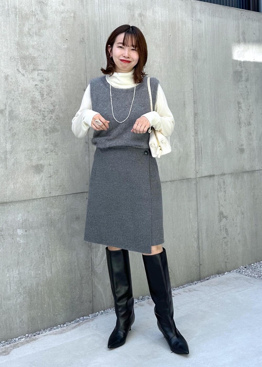 ブラック系のブーツ、ホワイト系のハンドバッグ、グレー系のベストを着用したレディースの秋コーディネートの2枚目の写真