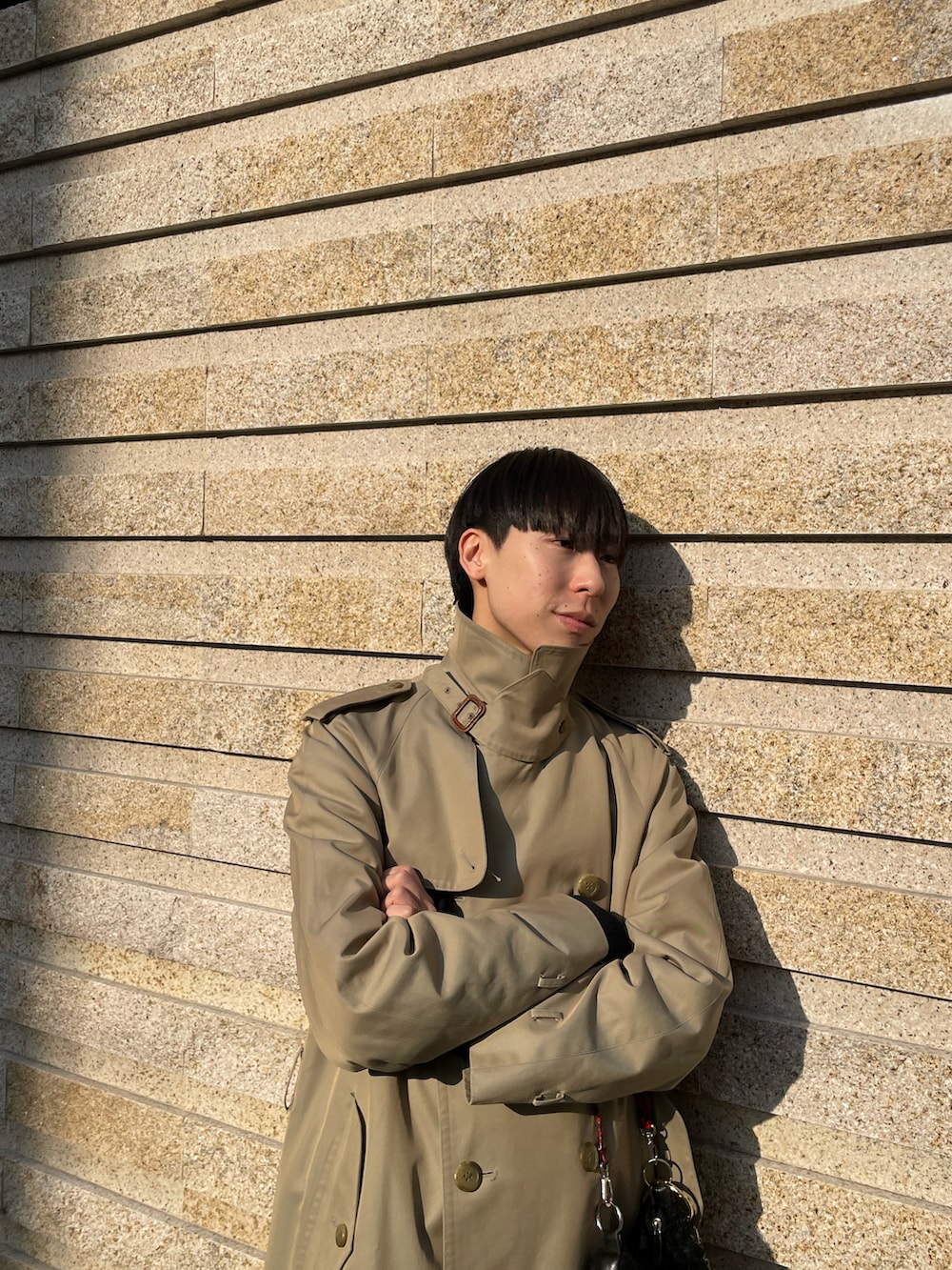 ブラック系のハンドバッグ、ベージュ系のトレンチコート、グレー系のスラックスを着用したメンズの春コーディネートの3枚目の写真
