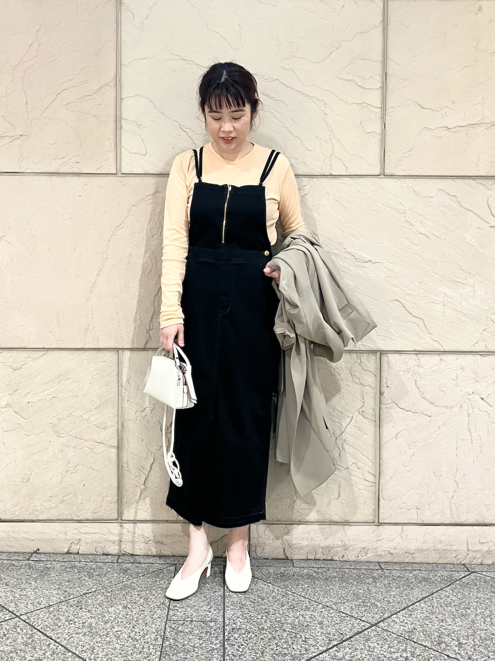 ベージュ系のトレンチコート、ブラック系のジャンパースカート、オレンジ系のTシャツ/カットソーを着用したレディースの春コーディネートの2枚目の写真