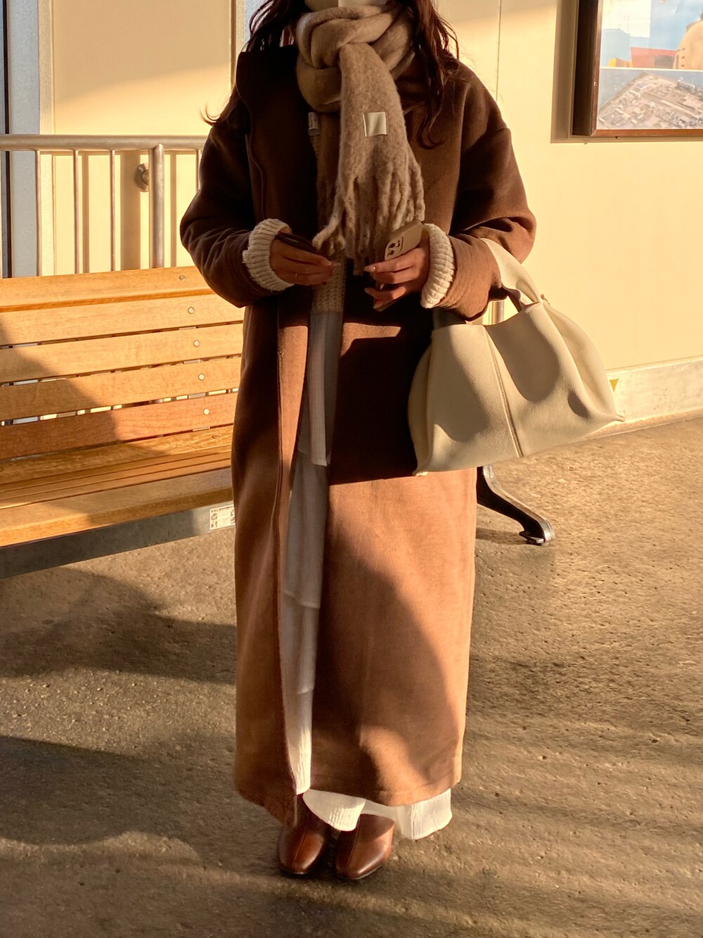 ブラウン系のノーカラーコート、ブラウン系のブーツ、ホワイト系のハンドバッグを着用したレディースの冬コーディネートの2枚目の写真