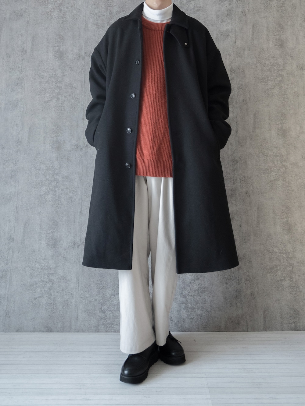 ブラック系のステンカラーコート、オレンジ系のニット/セーター、ホワイト系のTシャツ/カットソーを着用したメンズの冬コーディネートの2枚目の写真