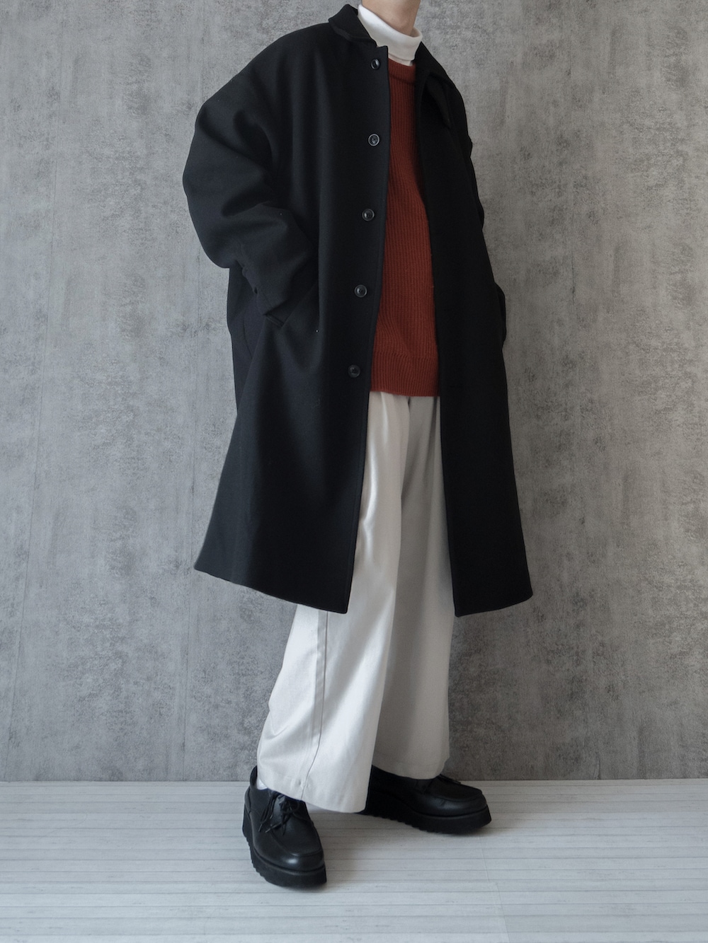 ブラック系のステンカラーコート、オレンジ系のニット/セーター、ホワイト系のTシャツ/カットソーを着用したメンズの冬コーディネートの3枚目の写真