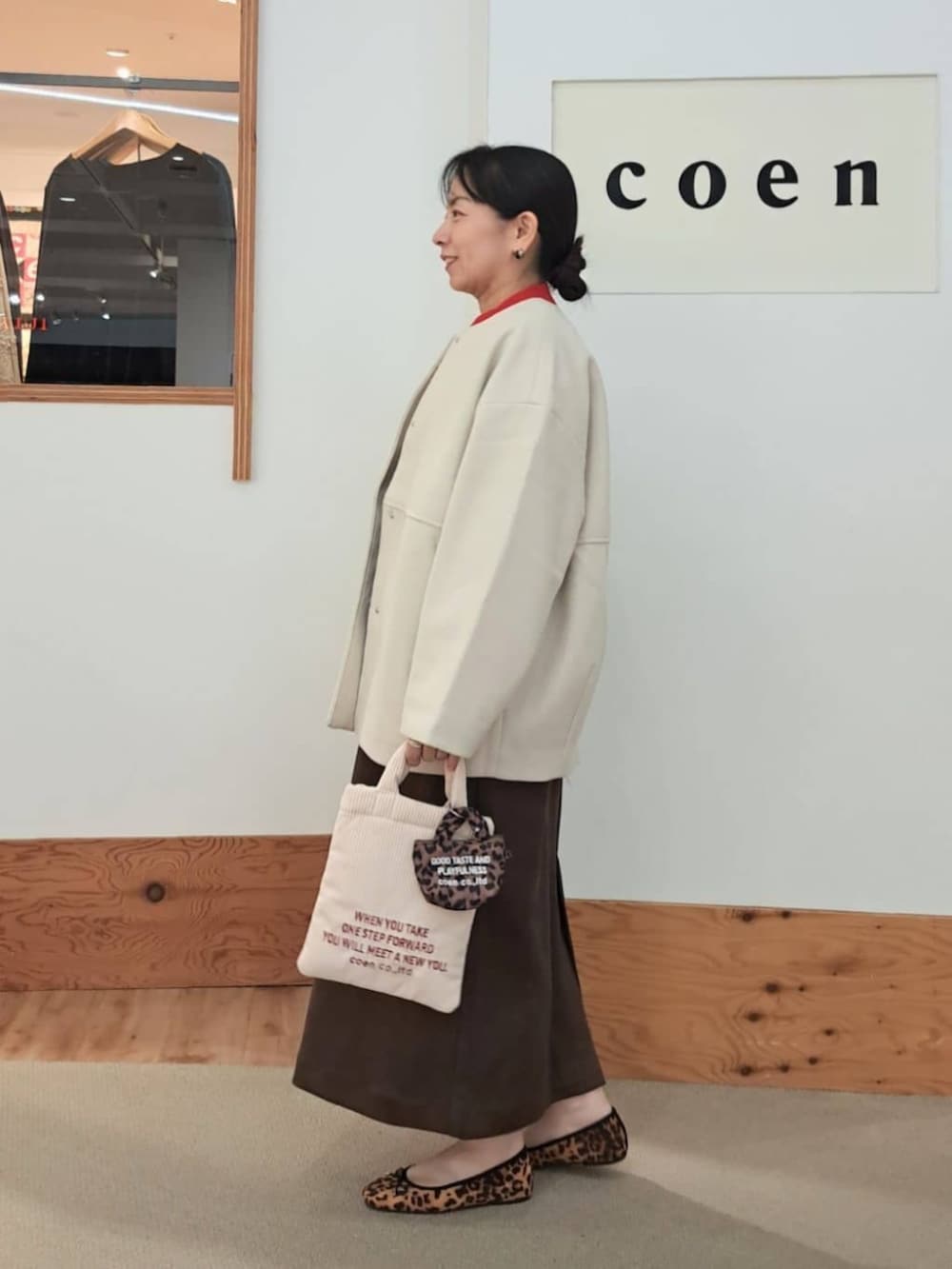 ベージュ系のノーカラーコート、ブラウン系のジャンパースカート、ベージュ系のショルダーバッグを着用したレディースの秋コーディネートの2枚目の写真