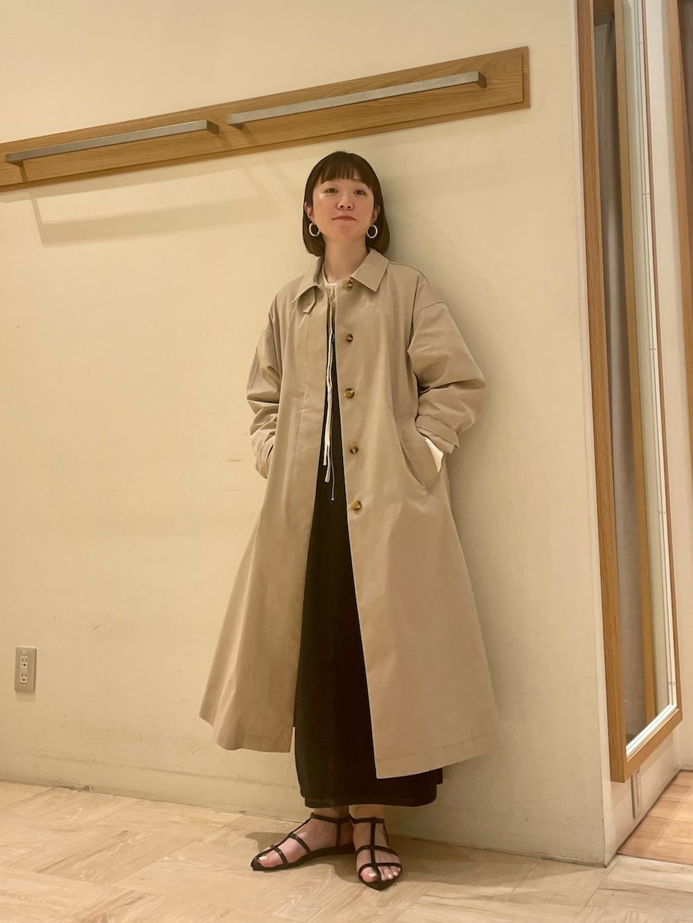 ベージュ系のステンカラーコート、ブラック系のジャンパースカート、ホワイト系のシャツ/ブラウスを着用したレディースの春コーディネートの2枚目の写真